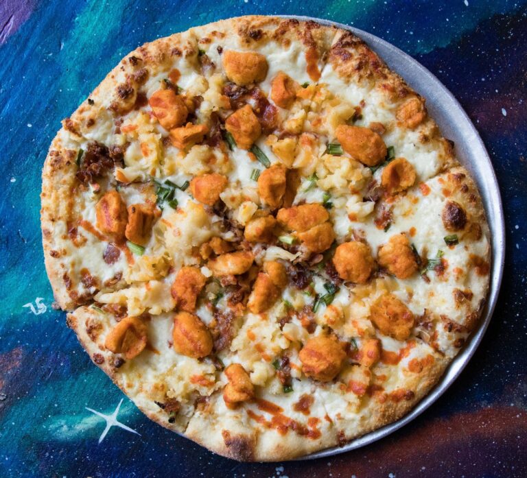 Specialty pizza topped with golden-brown chunks, served over a starry, space-printed table at Flying Saucer Pizza Company