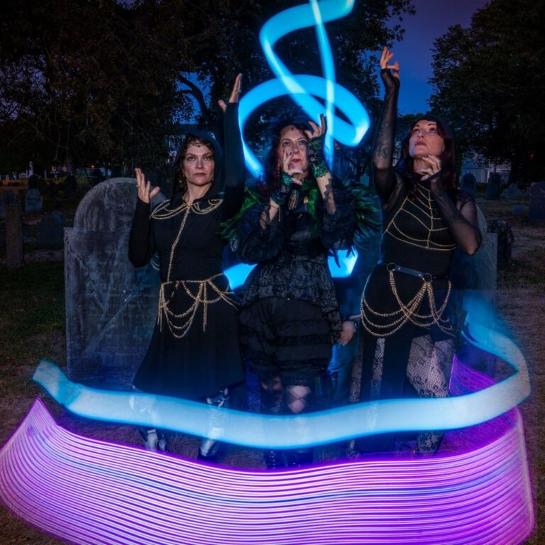 Three people in dark costumes among old headstones, circled by blue and purple light-painting trails